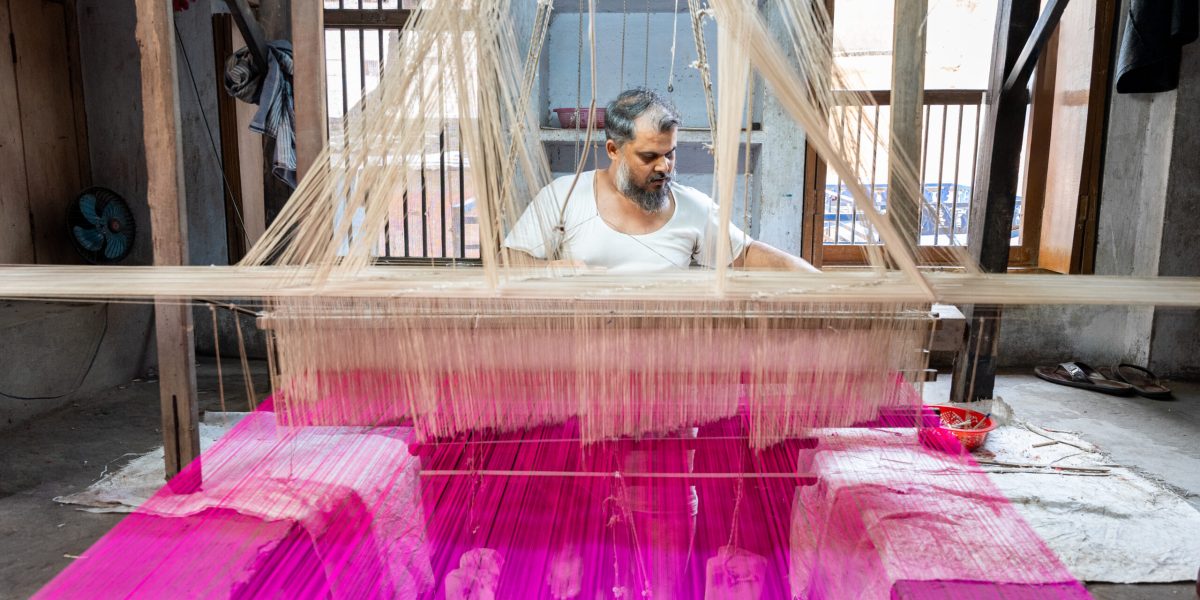 Indian man sitting at handloom weaving with purple thread Frontal view of an Indian man sitting in front of a handloom weaving with purple thread